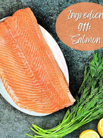 Large piece of salmon on a plate with fresh dill and lemon halves.