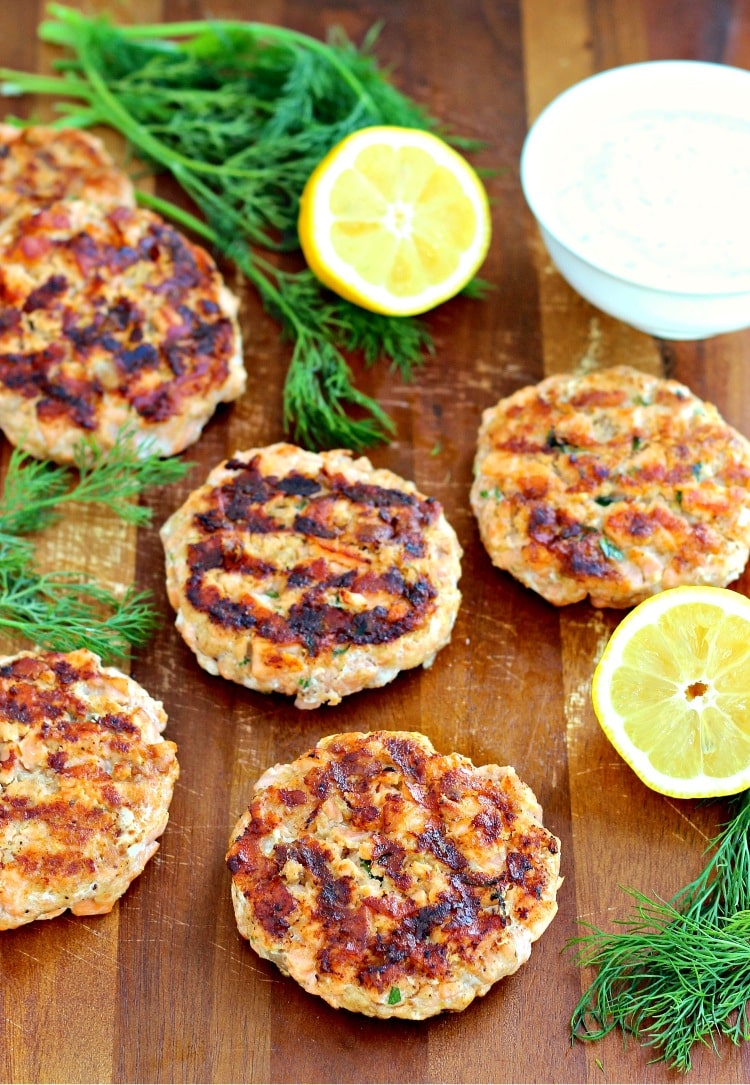 Salmon Burgers on a wooden board with lemon halves and dill sprigs.