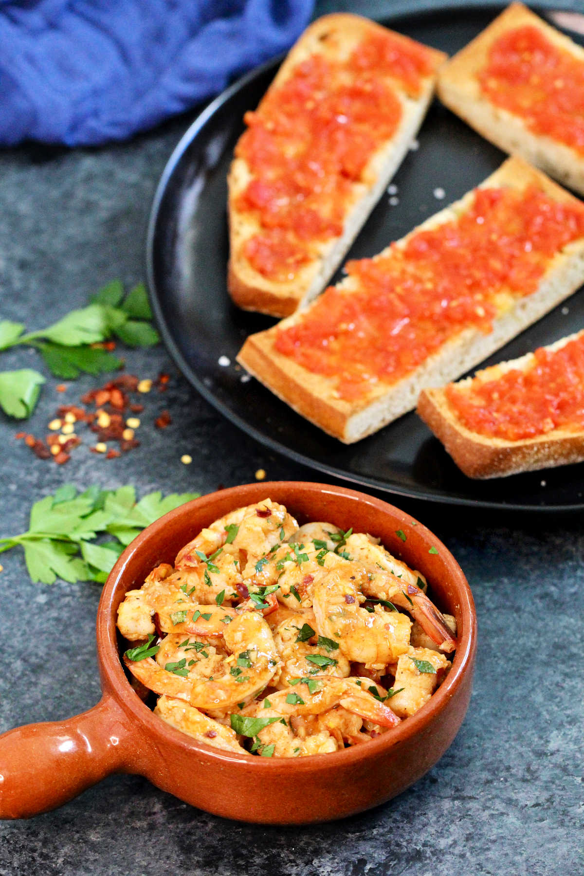 Overhead shot of Spanish garlic shrimp in a brown dish with pan con tomate in the background.