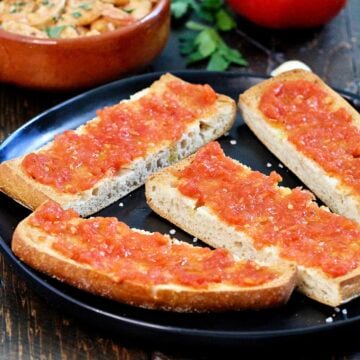 Pan con tomate on a black plate with Spanish garlic shrimp and a tomato in the background.