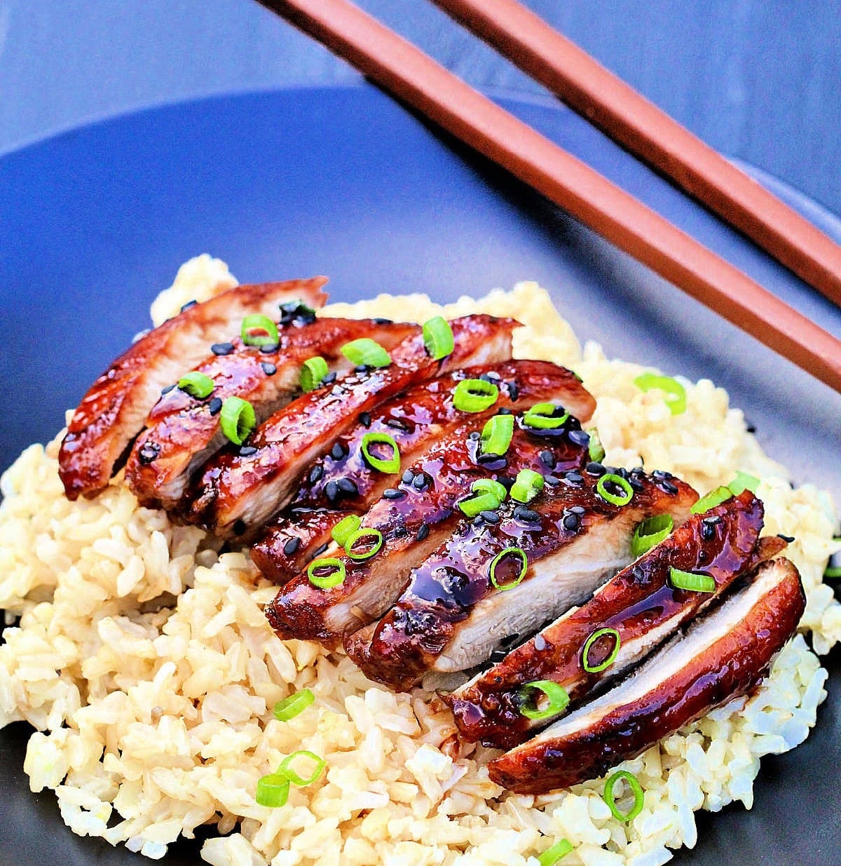 Sliced chicken teriyaki on a bed of brown rice with brown chopsticks nearby.