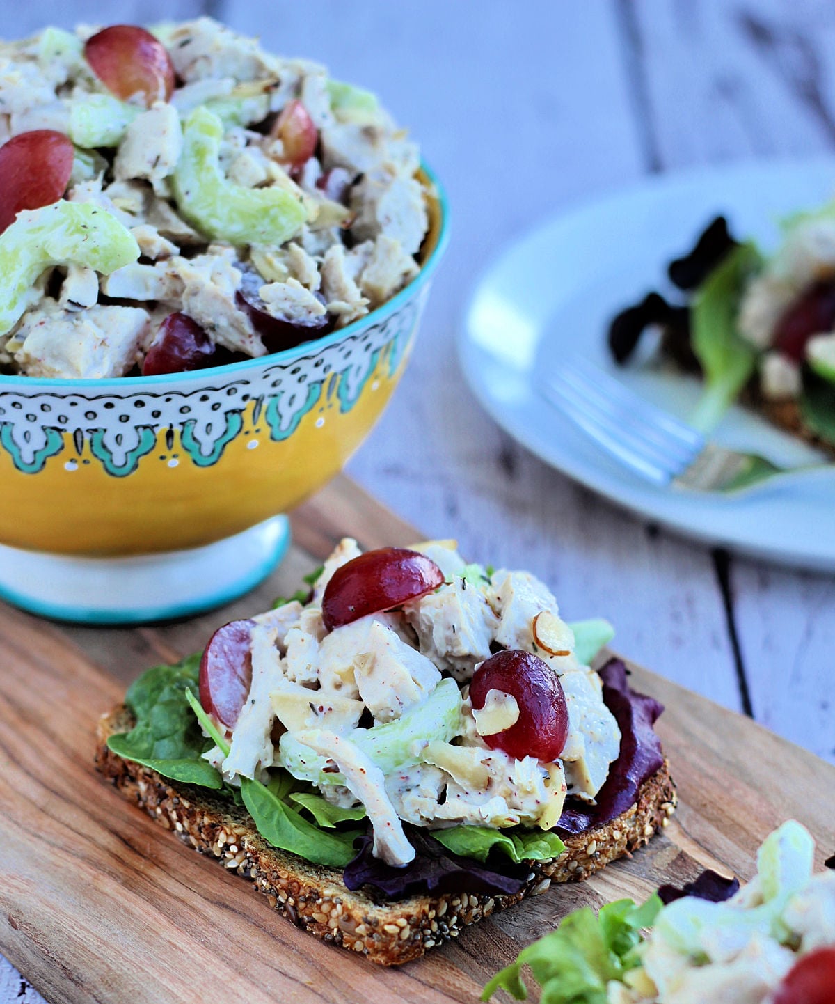 Chicken salad with grapes and almonds on a slice of multigrain bread with a bowl of chicken salad in the background.