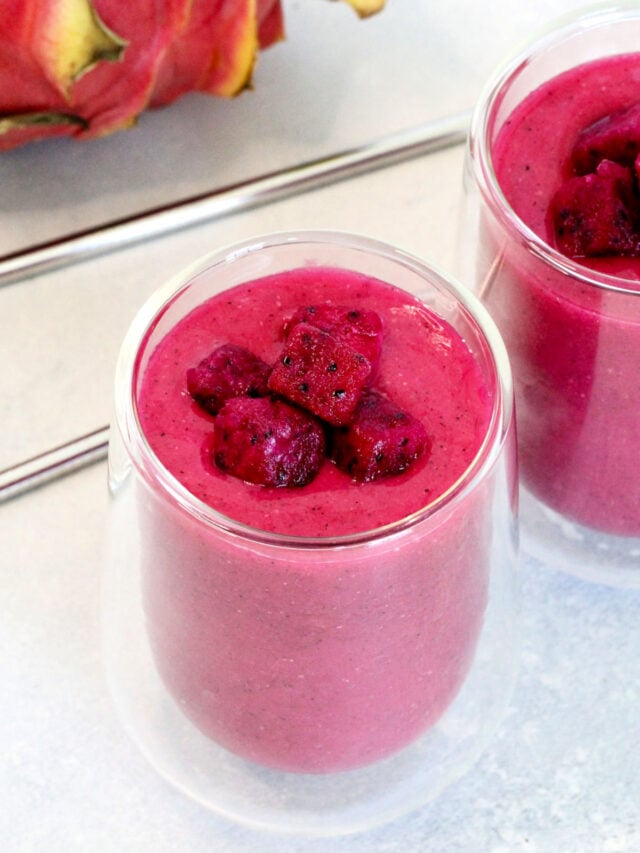 Two dragon fruit smoothies in clear glasses with metal straws and a dragon fruit in the background.