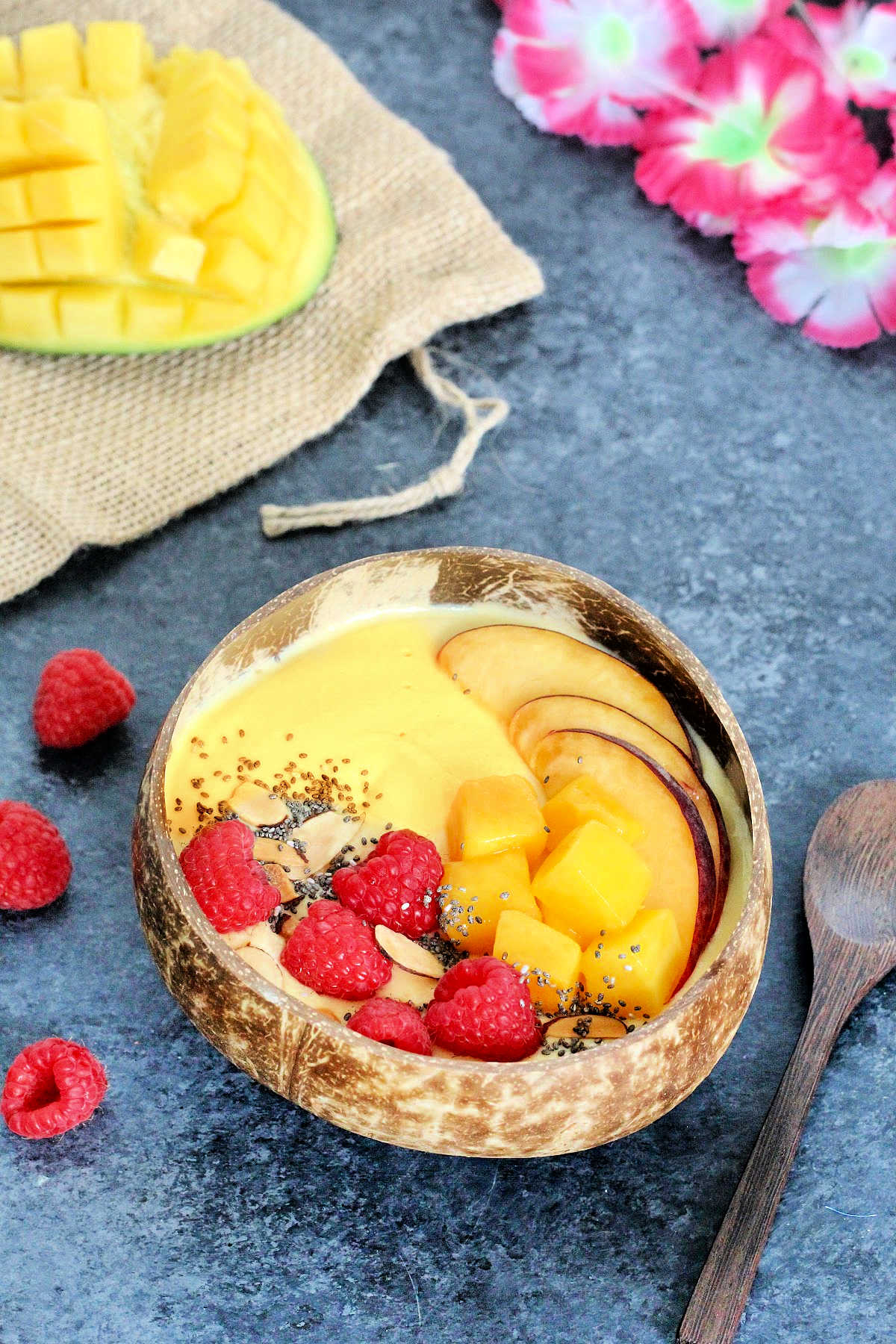 A Mango Smoothie Bowl in a wooden bowl surrounded by a wooden spoon, a cut mango, raspberries and pink flowers.