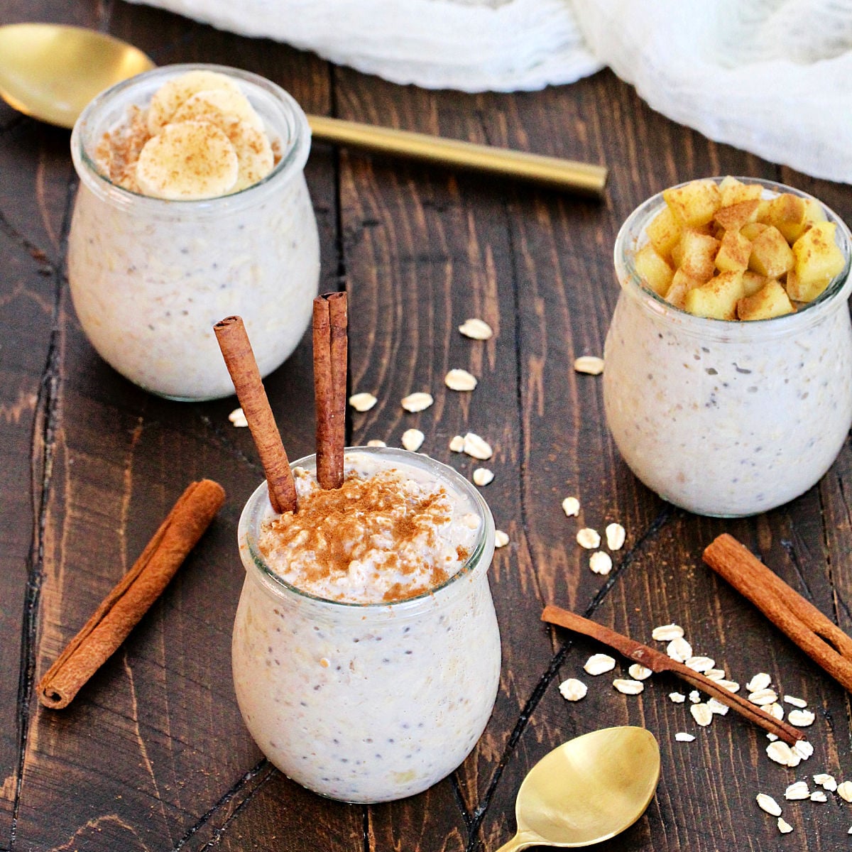 3 jars of Cinnamon Overnight Oats in glass jars with different toppings surrounded by cinnamon sticks, oats and gold spoons.