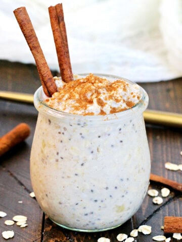Cinnamon Overnight Oats in a glass jar topped with cinnamon sticks with a gold spoon and oats lying around it.