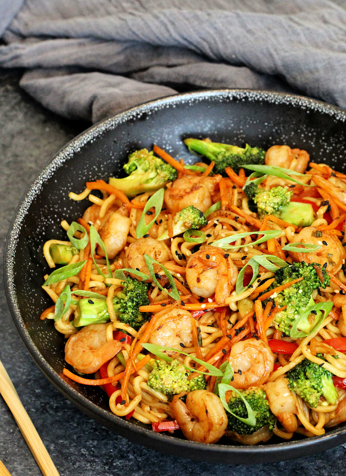 Shrimp Stir Fry with Noodles in a black bowl with a gray napkin and wooden chopsticks nearby.