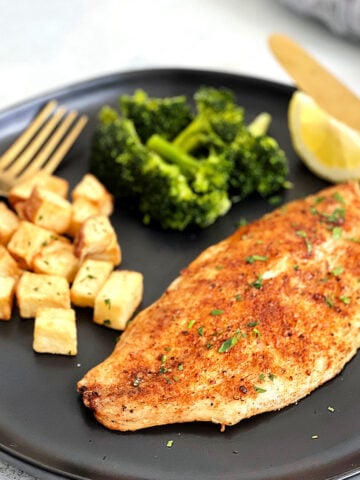 Air fryer tilapia on a black plate with diced potatoes, broccoli and a lemon wedge.