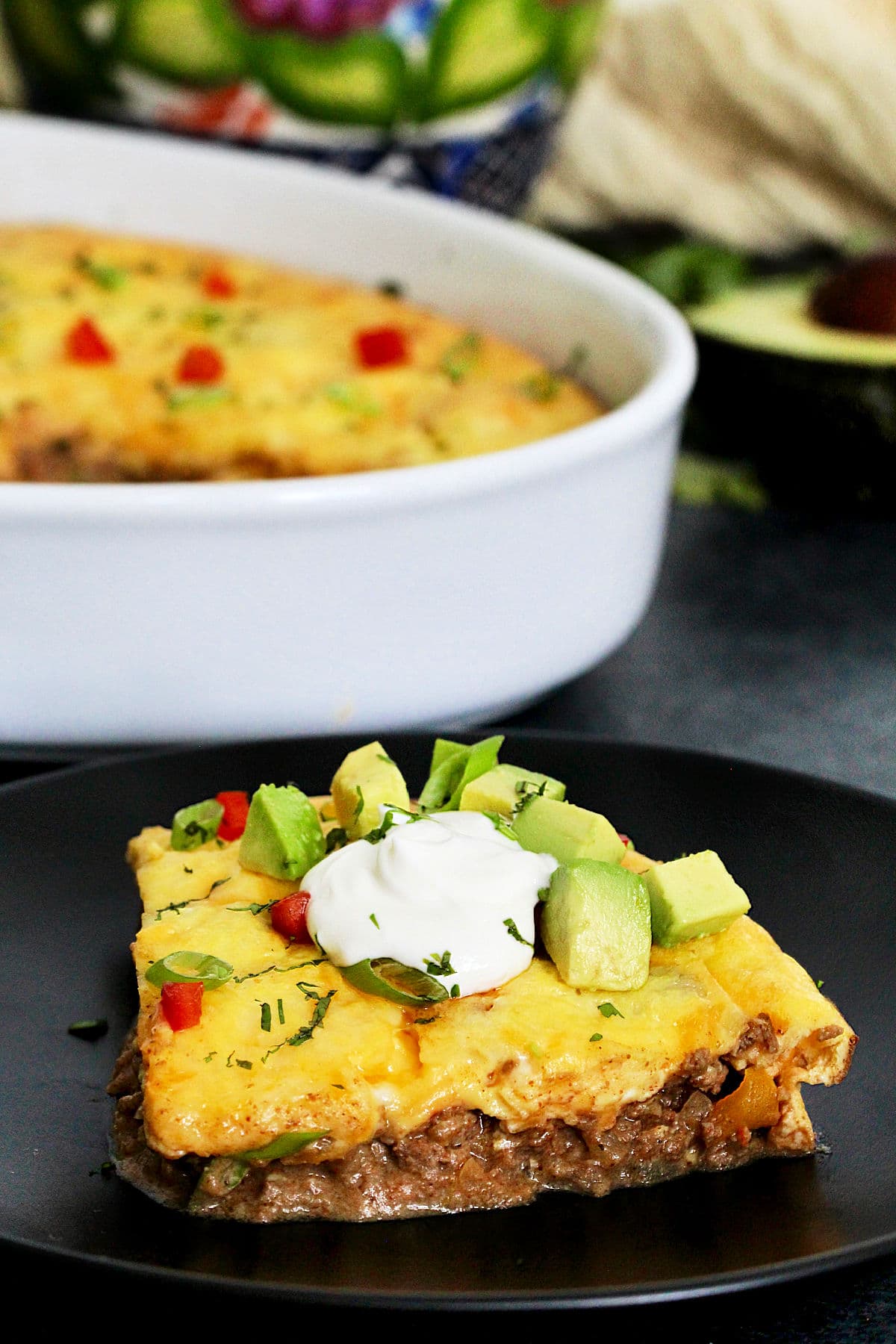Piece of easy keto taco casserole on a black plate in front of a white casserole dish.