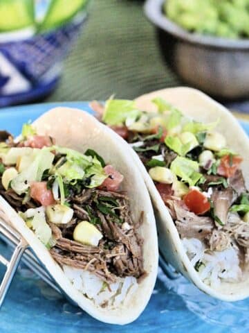 One barbacoa taco and one carnitas taco in a metal taco stand with guacamole in background.