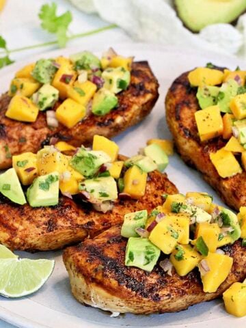 Grilled chicken with mango avocado salsa on a white plate with mango and avocado in the background.