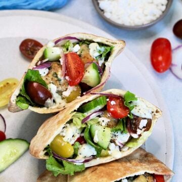 Three Greek pita sandwiches on a round plate with a bowl of feta cheese next to it.