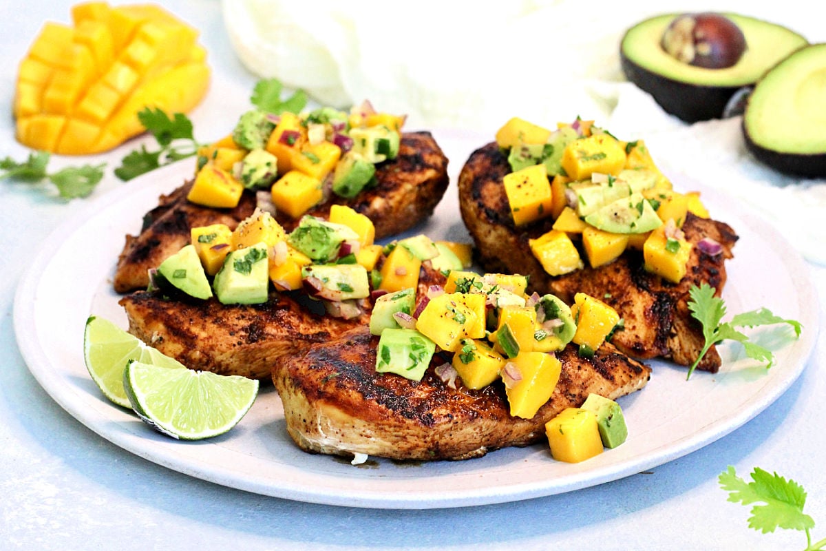 Grilled chicken with mango avocado salsa on a white plate with mango and avocado in the background.