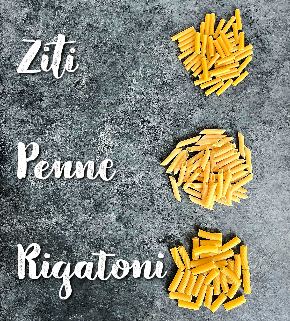 Different types of pasta on a gray board labeled "Ziti", "Penne" and "Rigatoni"