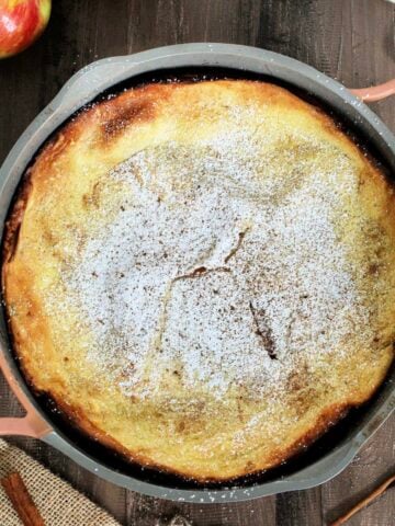 An Apple Dutch Baby in a pan on a wooden board surrounded by apples, cinnamon sticks and measuring spoons.