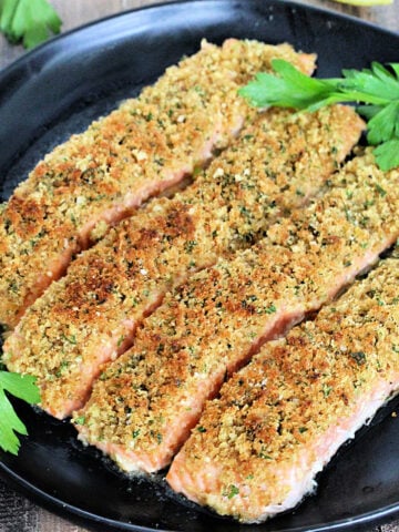 Four pieces of salmon oreganata on a black plate with parsley and lemons.