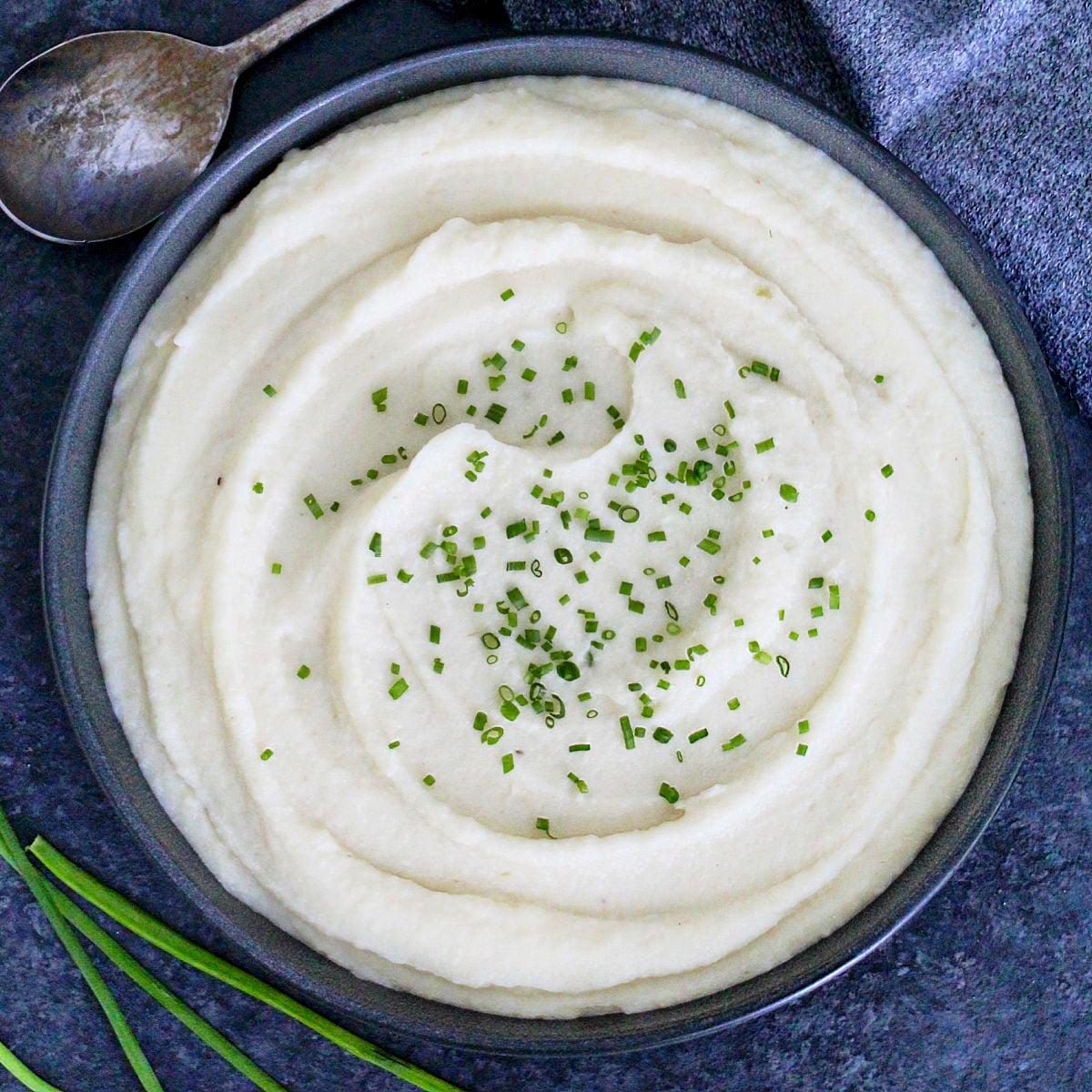 Celeriac (celery root) puree garnished with scallions in a dark bowl with a metal spoon and scallions around it.