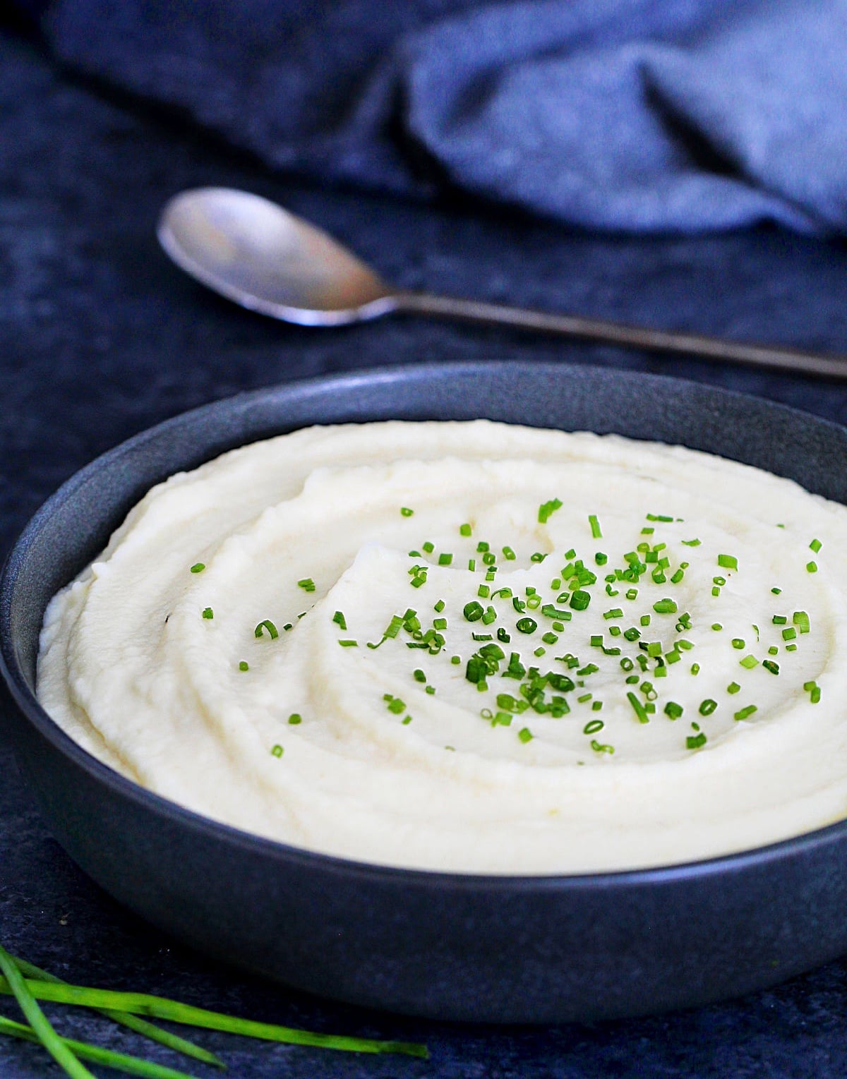 Celeriac (celery root) puree garnished with scallions in a dark bowl with a metal spoon and scallions around it.
