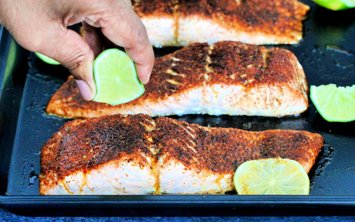 Lime being squeezed on salmon fillets on a black baking sheet.