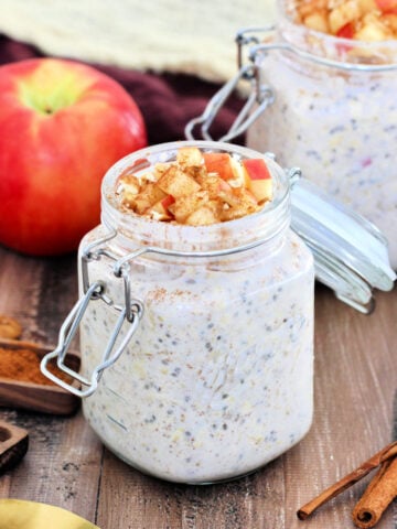 Two jars of apple cinnamon overnight oats.surrounded by an apple, cinnamon sticks, brown napkins and wooden measuring spoons.