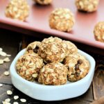 A stack of 3-Ingredient Peanut Butter Oatmeal Balls on a white dish in front of more balls on a pink baking sheet.