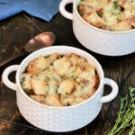 Two bowls of healthy French onion soup on a wooden board with thyme ,a blue napkin and a bronze spoon nearby.