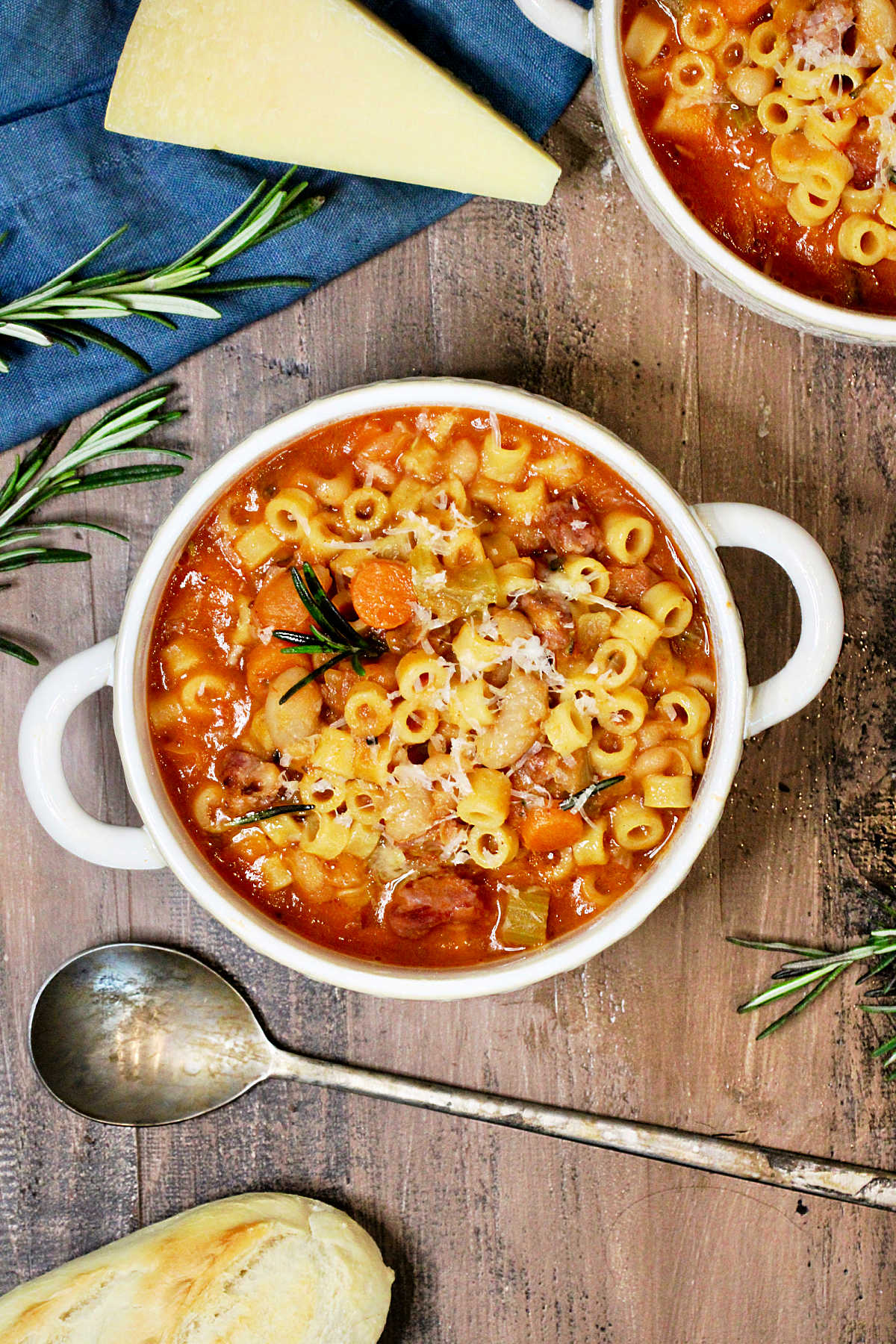 A bowl of pasta e fagioli soup on a wooden board surrounded by parmesan cheese, rosemary and a blue napkin.