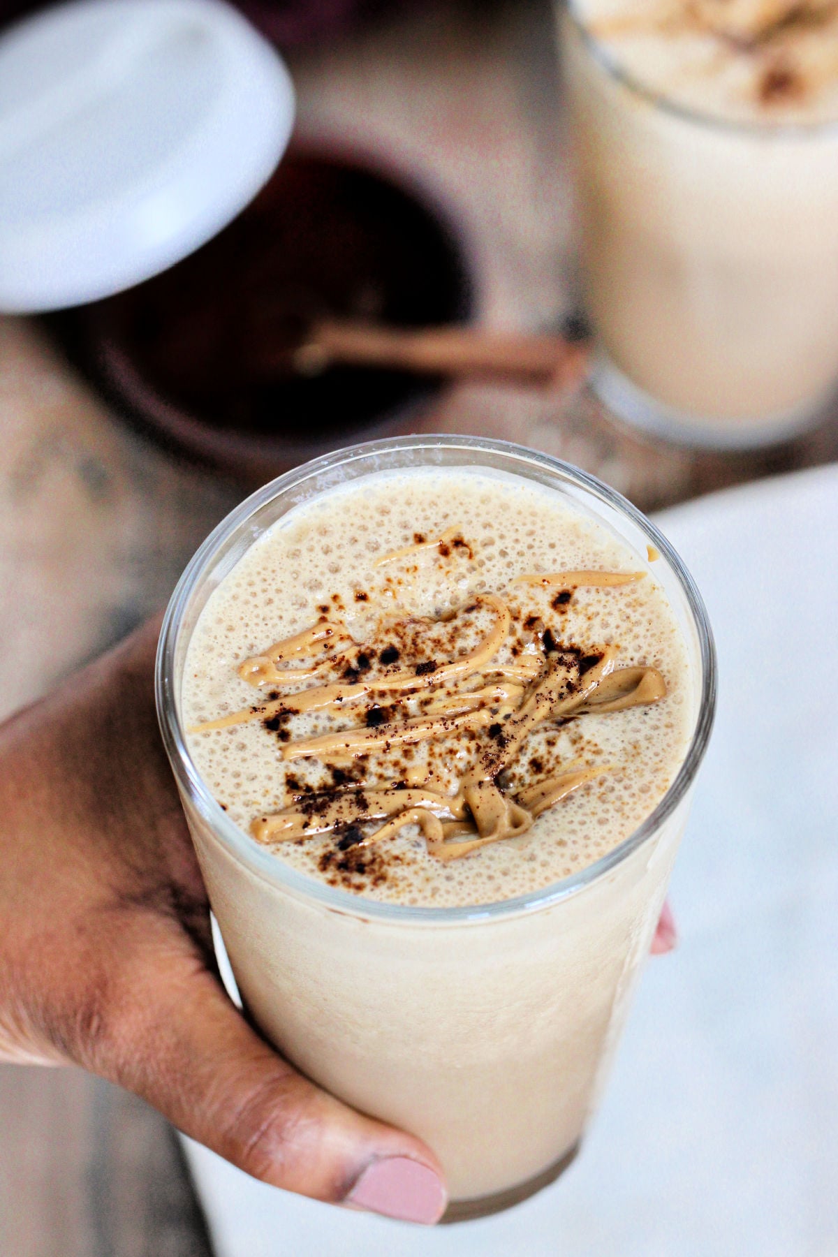 A coffee protein smoothie being held above a wooden board with another smoothie in the background.