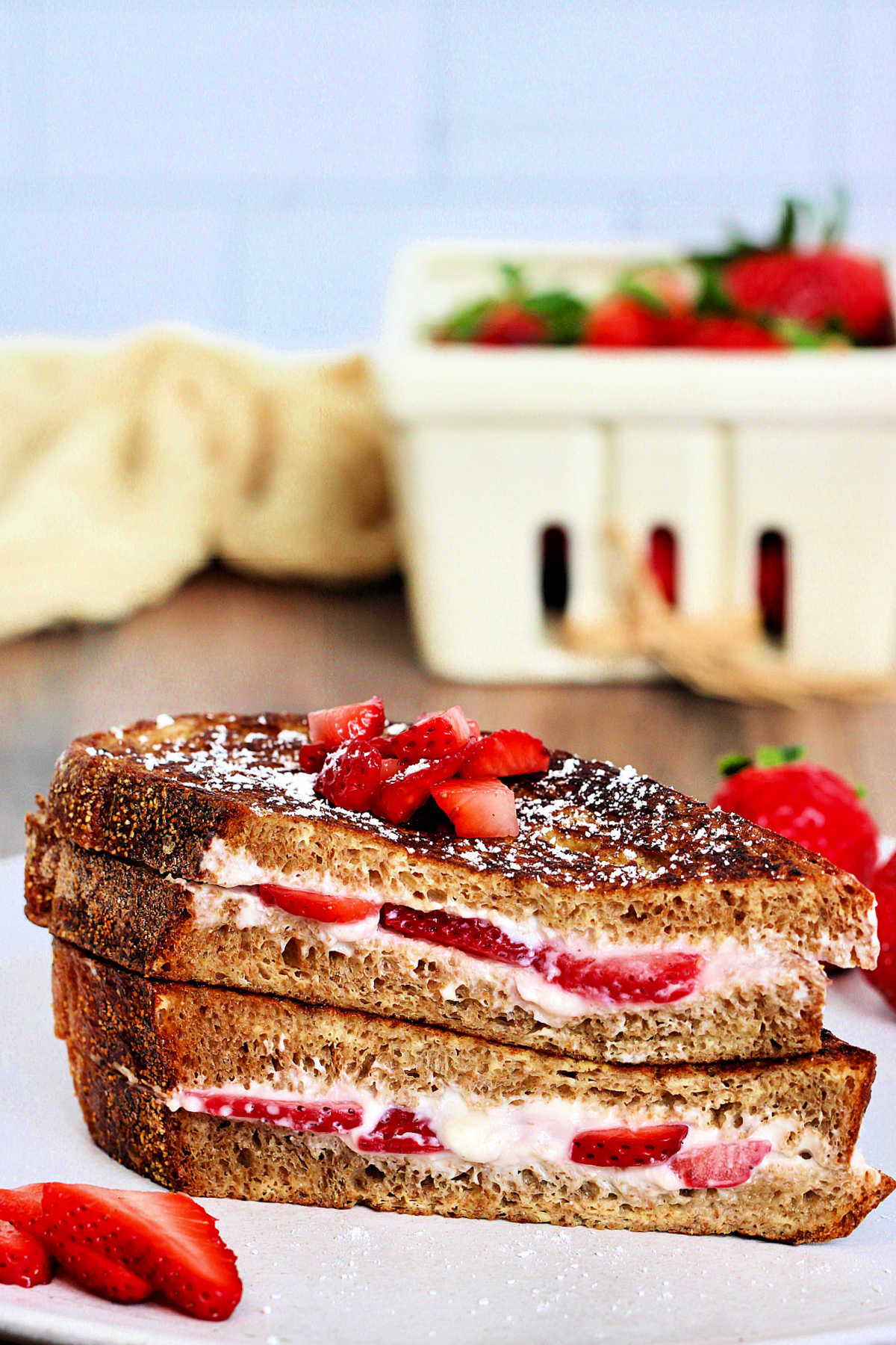 Two pieces of strawberry stuffed French toast stacked on a white plate with a container of strawberries in the background.