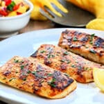 Grilled mahi mahi on a white plate with a bowl of mango salsa and a yellow napkin in the background.