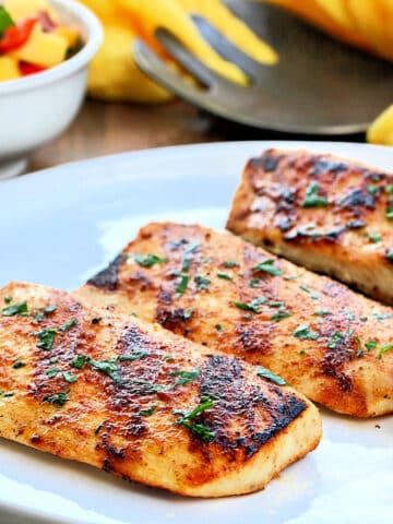 Grilled mahi mahi on a white plate with a bowl of mango salsa and a yellow napkin in the background.
