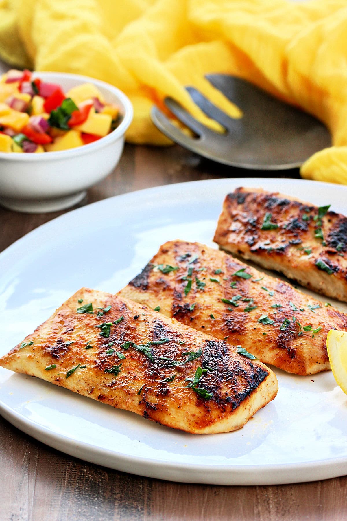 Grilled mahi mahi on a white plate with a bowl of mango salsa and a yellow napkin in the background.