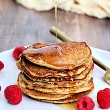 Maple syrup being poured on a stack of 3-Ingredient banana protein pancakes on a white plate with raspberries and a gold folk around them.