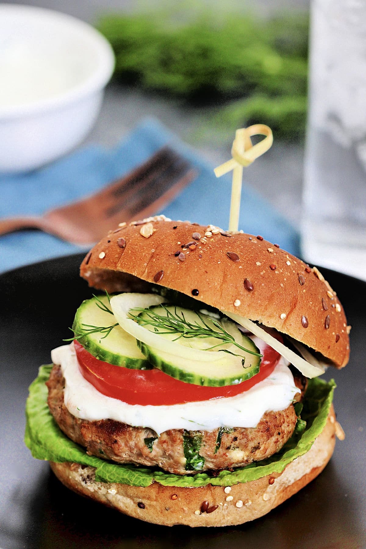 Greek turkey burger on a black plate with a blue napkin, fork and herbs in the background.