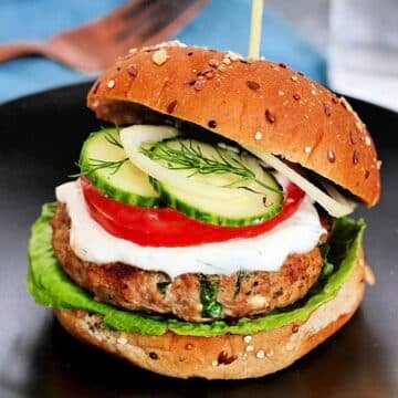 Greek turkey burger on a black plate with a blue napkin and a fork in the background.