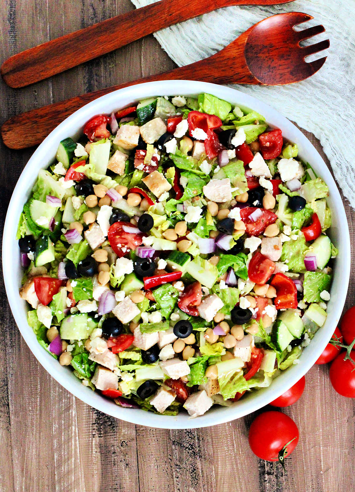 Mediterranean chopped salad in a white bowl with tomoatoes and woodern utensils nearby.