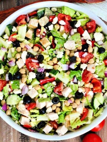 Mediterranean chopped salad in a white bowl with tomoatoes and woodern utensils nearby.
