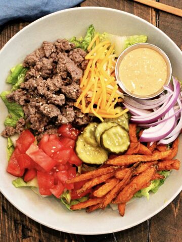 A burger bowl on a wooden board with a blue napkin and bronze fork nearby.