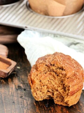 Easy Pumpkin Banana Muffin with a bite out of it near a muffin pan and wooden measuring spoons.