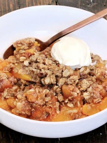 Gluten free peach crisp in a white bowl on a wooden board with a scoop of vanilla ice cream in the bowl.