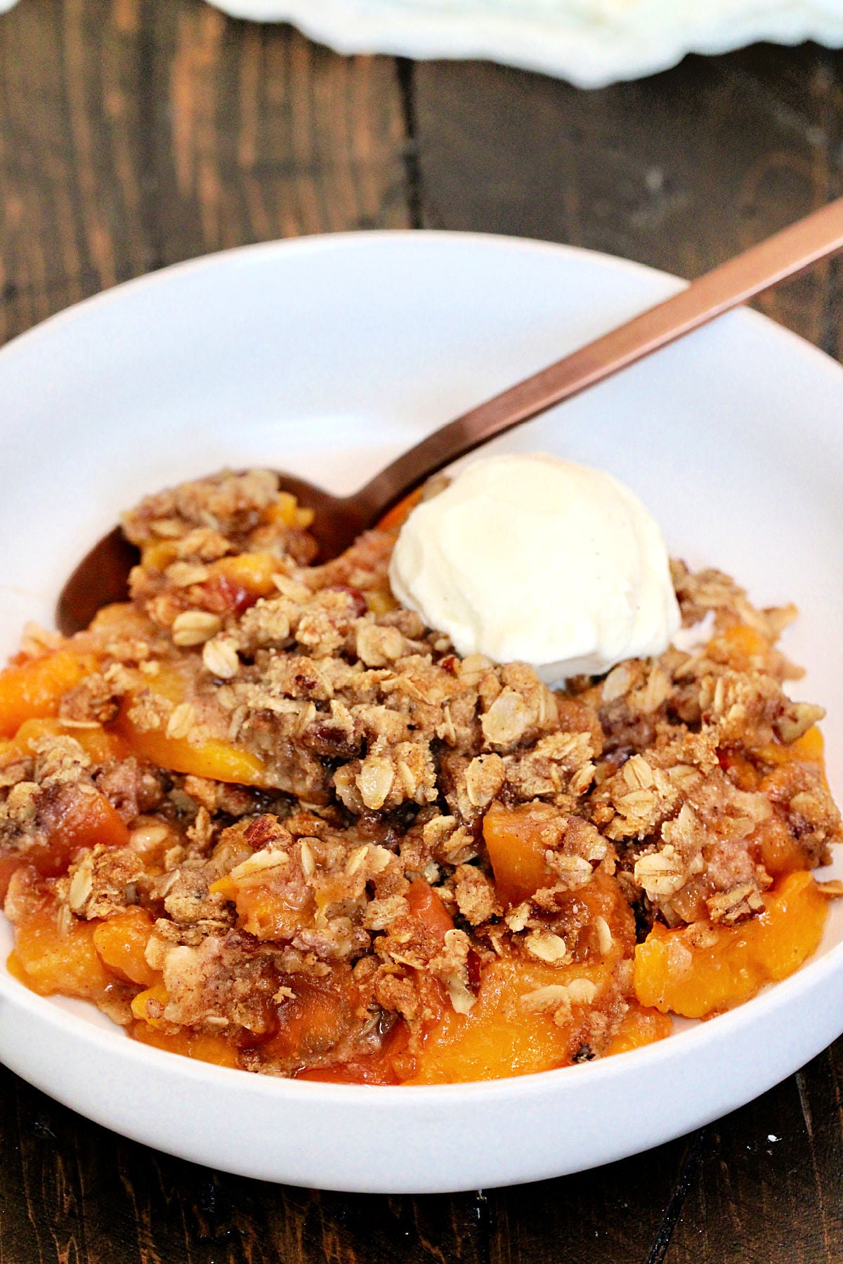 Gluten free peach crisp in a white bowl on a wooden board with a scoop of vanilla ice cream in the bowl.
